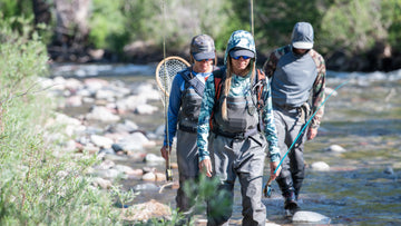 Three people in fishing gear walking along a riverbank.